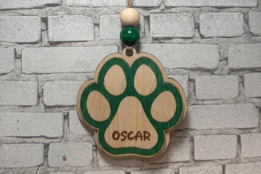 A personalized wooden paw print ornament hanging from a string against a white brick wall. The ornament is light wood with a green paw print design, the name 'OSCAR' engraved in the pad, and a green bead accent below a cream-colored bead.