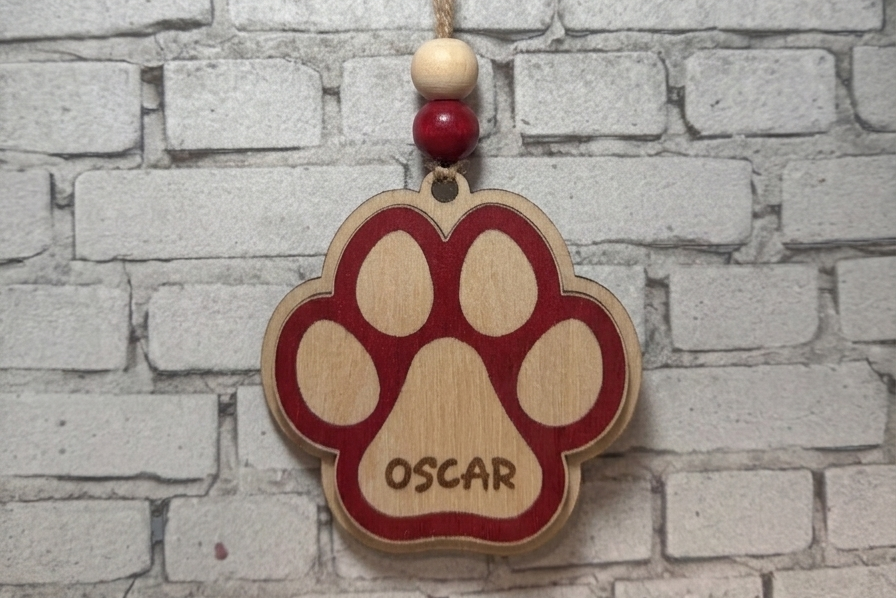 A personalized wooden paw print ornament hanging from a string with two beads, featuring the name 'OSCAR' engraved in the center, set against a white brick wall background.