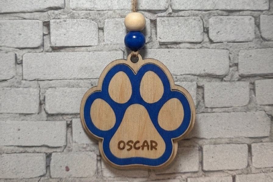 A personalized wooden paw print hanging ornament engraved with the name 'OSCAR', featuring blue painted details and a natural wood finish, hanging against a white brick wall background.