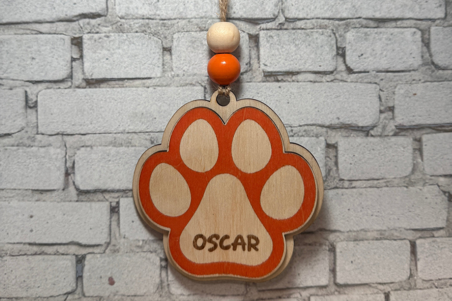 A wooden paw print hanging ornament engraved with the name 'OSCAR', hanging from a string against a white brick wall background.