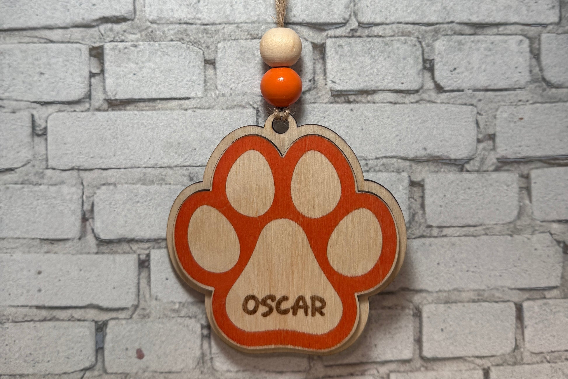 A wooden paw print hanging ornament engraved with the name 'OSCAR', hanging from a string against a white brick wall background.